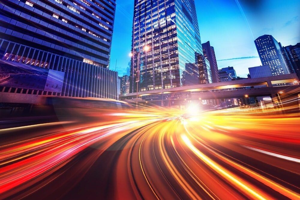 Long-exposure light trails streak through a modern city street lined with illuminated skyscrapers at twilight.