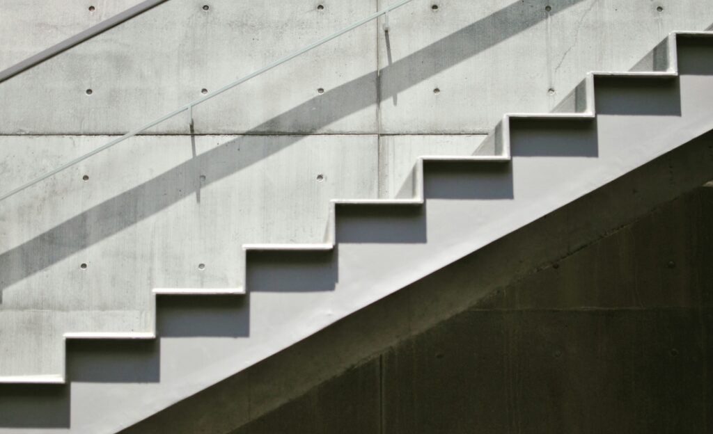 Modern concrete staircase ascending diagonally against a smooth concrete wall with a glass railing casting diagonal shadows.
