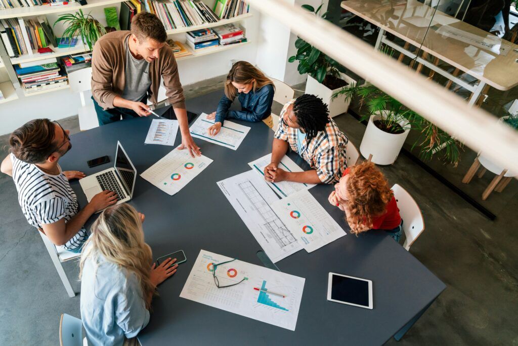 Diverse team of six professionals collaborating around a table with charts, reports, and laptops in a modern office.