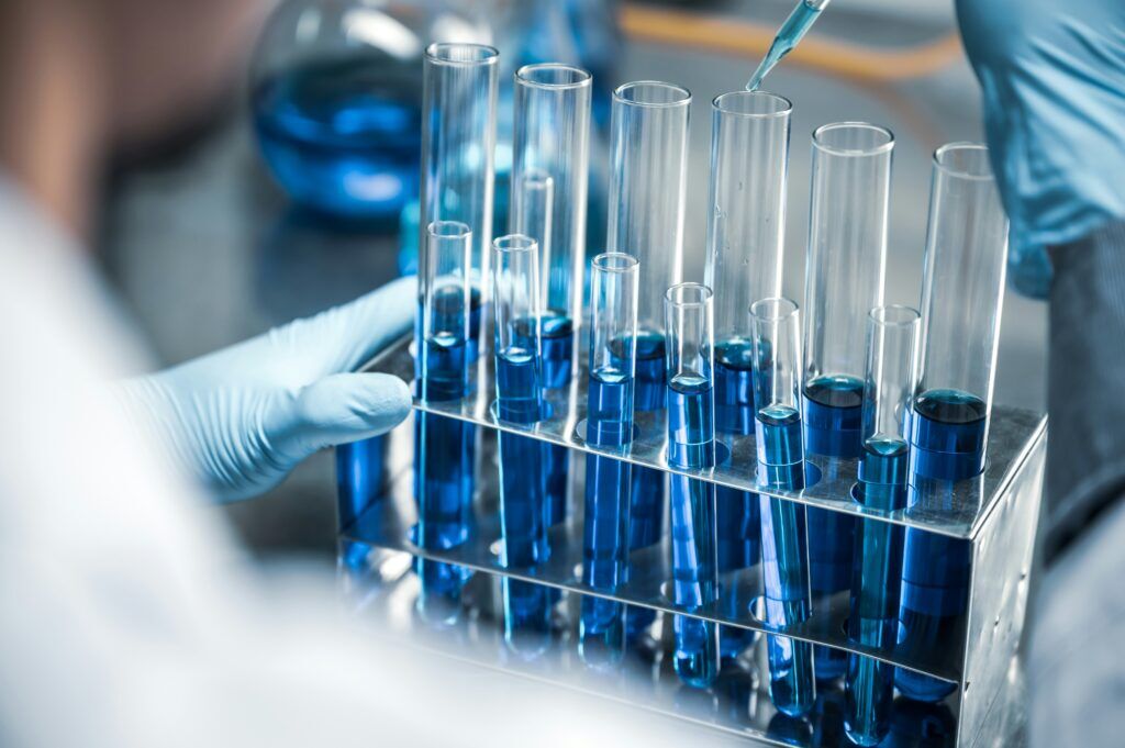 Gloved hands working with test tubes filled with blue liquid in a metal rack while a pipette dispenses solution above.