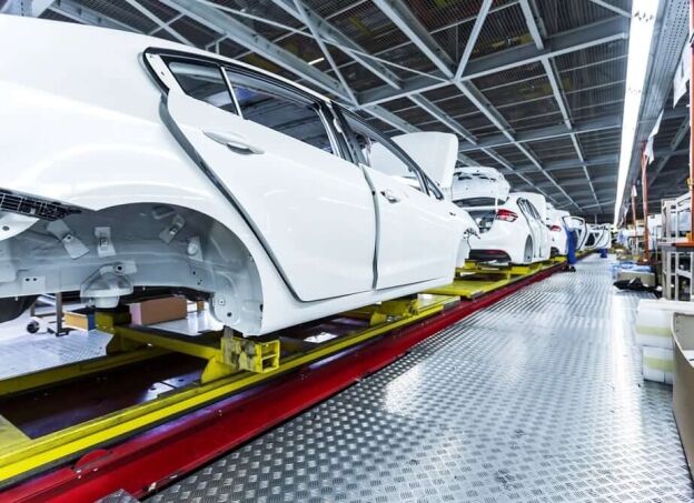 White car bodies move along a factory assembly line on yellow conveyor platforms inside an automotive manufacturing plant.