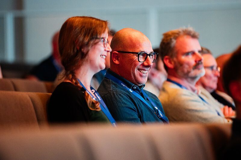 Smiling conference attendees wearing lanyards sit in auditorium seats, engaged in a presentation.