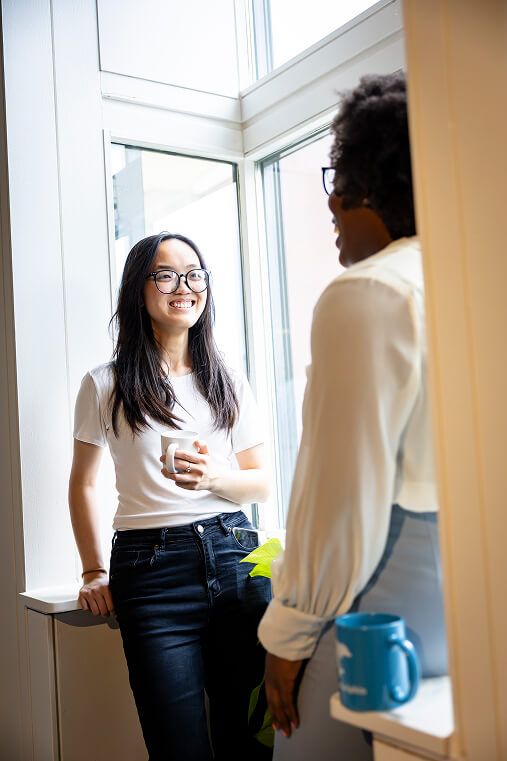 Two women holding coffee mugs chat and smile by a sunlit office window during a casual break.