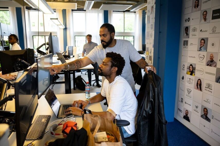 Two colleagues collaborating at a desk with laptops in a modern office with a client photo wall nearby.