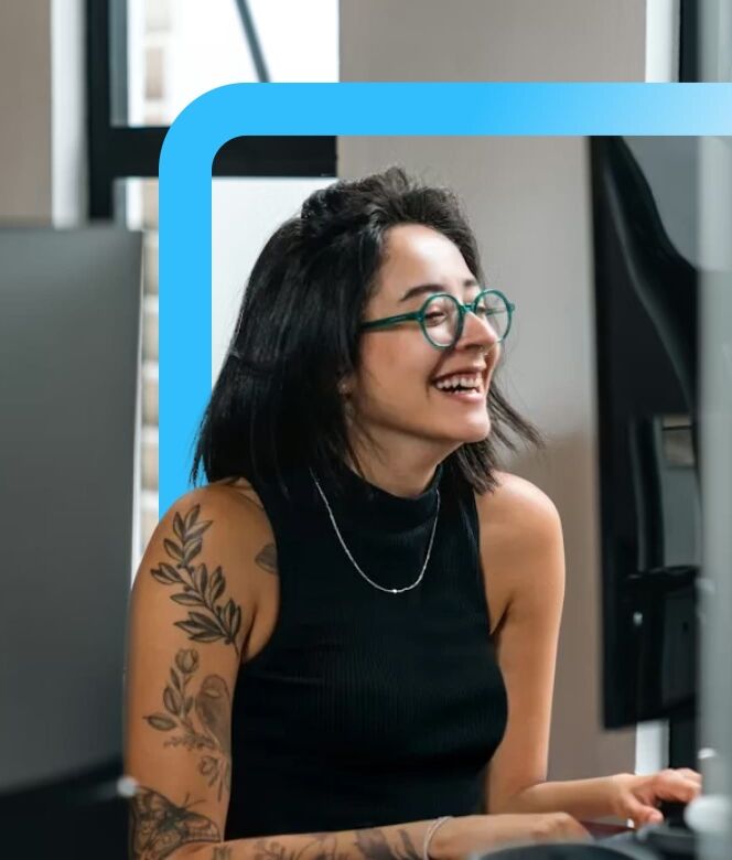 Smiling woman with green glasses and floral arm tattoos working at a desk with computer monitors.
