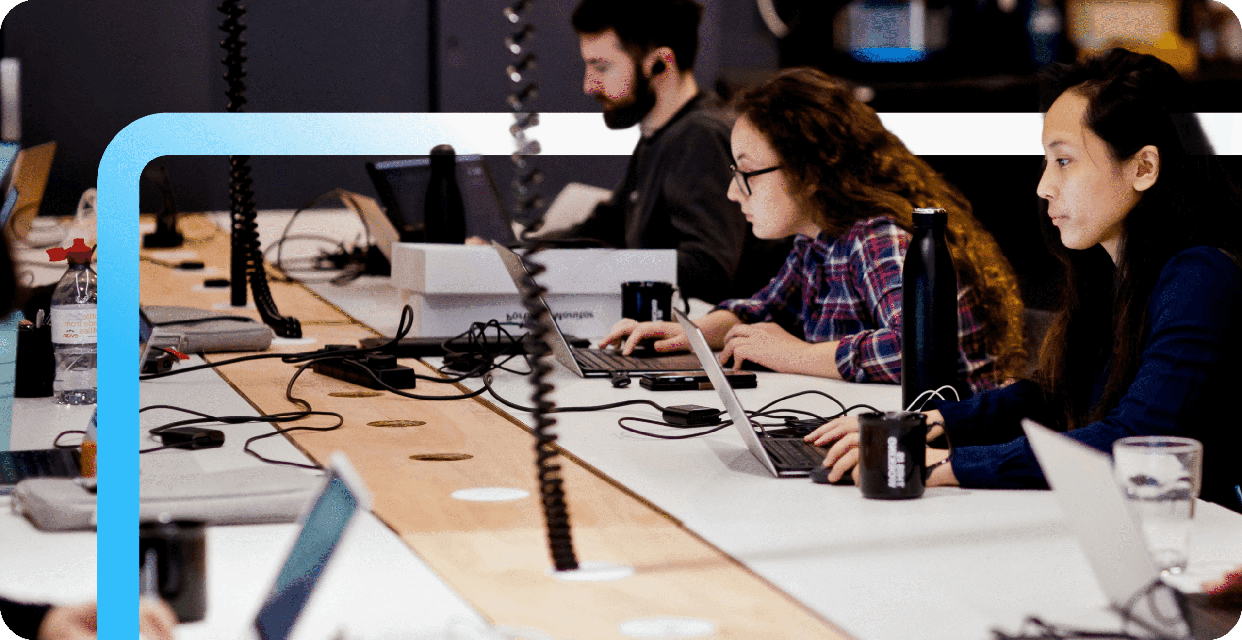 Diverse software developers working on laptops at a shared desk in a modern open-plan office with a blue accent border.