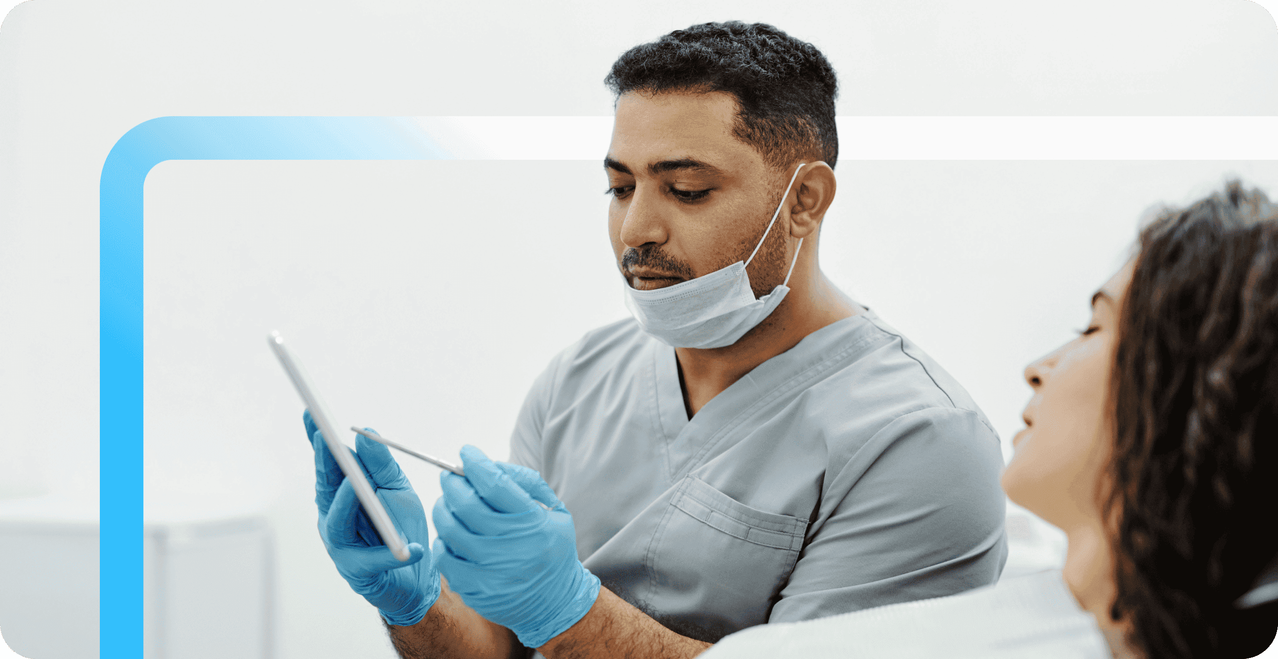 A dental professional in gray scrubs and blue gloves reviews a tablet while consulting with a female patient.
