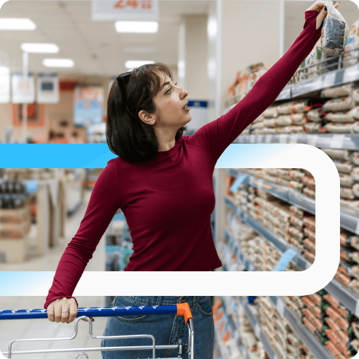 Woman in a burgundy top reaching for a product on a high shelf while pushing a shopping cart in a retail store aisle.