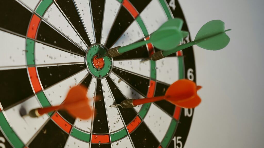 Green and orange darts stuck in a dartboard with one green dart hitting near the bullseye center.