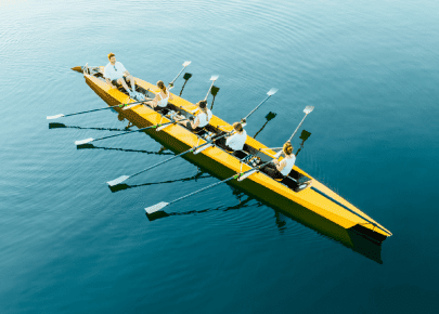 Four rowers paddling together in a yellow rowing boat on calm blue water, symbolizing teamwork and collaboration.