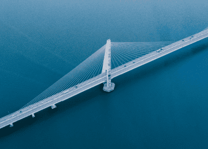 Aerial view of a cable-stayed bridge with a tall central pylon stretching diagonally across calm blue water.