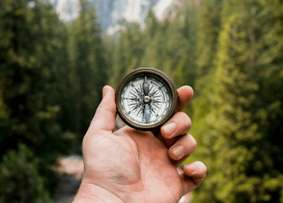 A hand holding a vintage compass against a blurred backdrop of evergreen trees and mountain peaks.