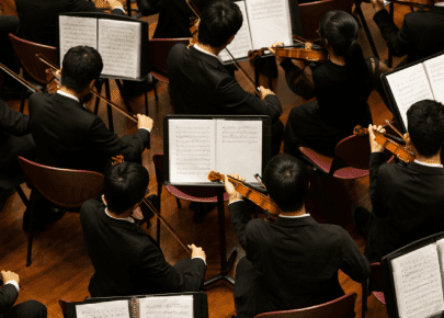 Overhead view of orchestra musicians in black attire playing violins while reading sheet music on stands.