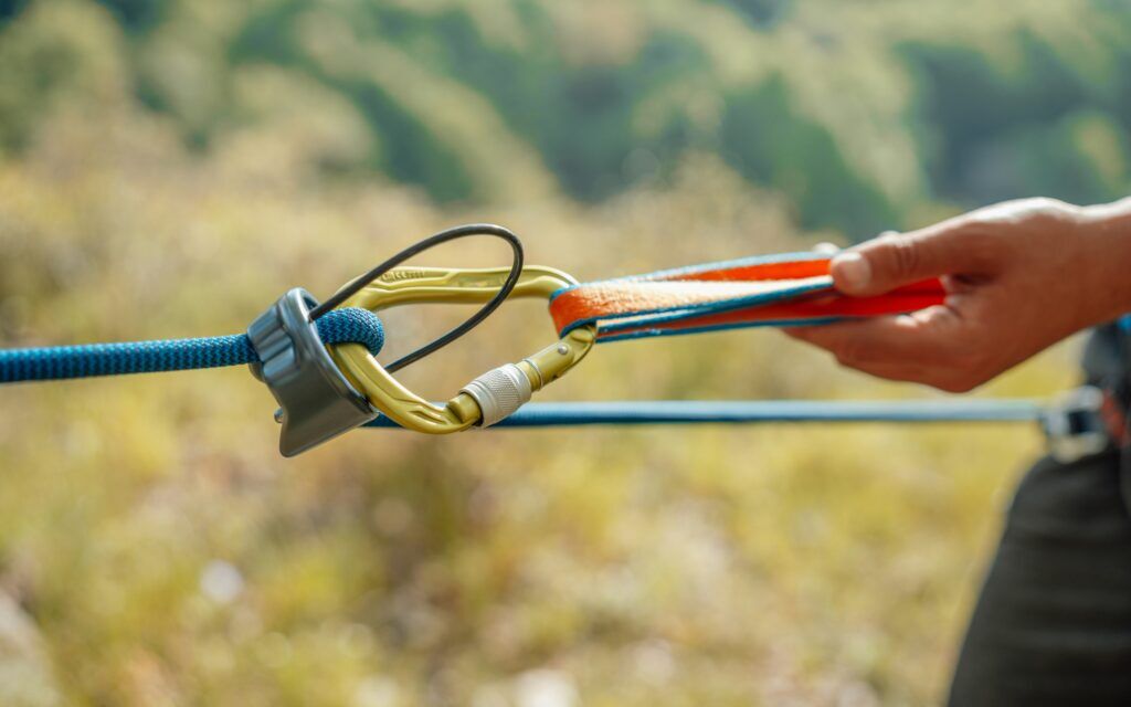 A hand gripping a carabiner and belay device connected to climbing ropes against a blurred mountain backdrop.