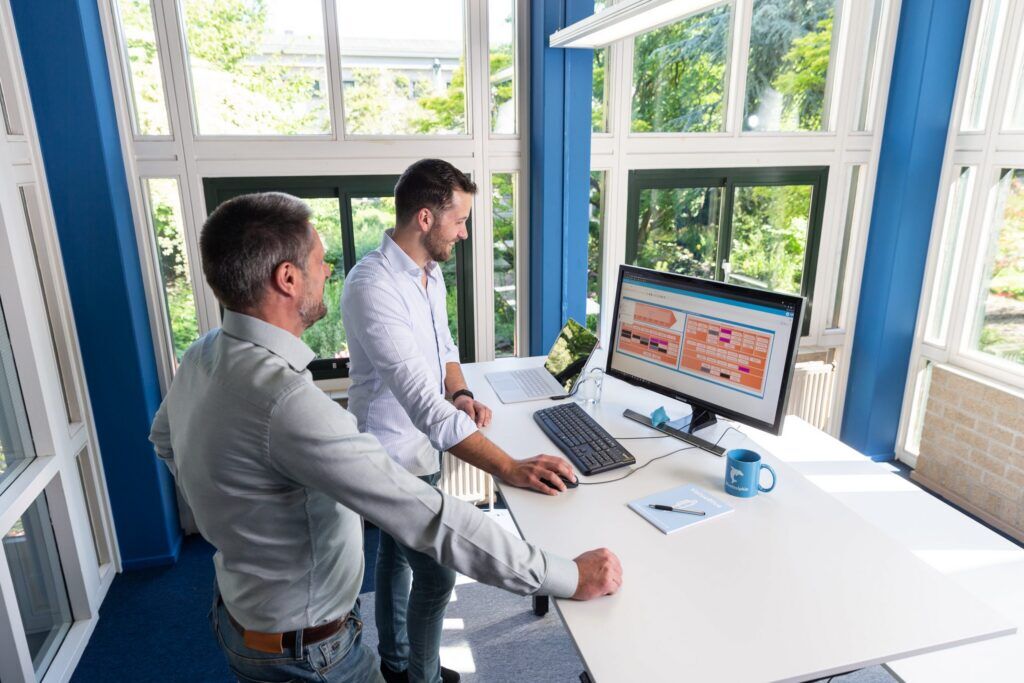 Two colleagues reviewing enterprise architecture diagrams on a monitor at a bright, blue-framed office desk.