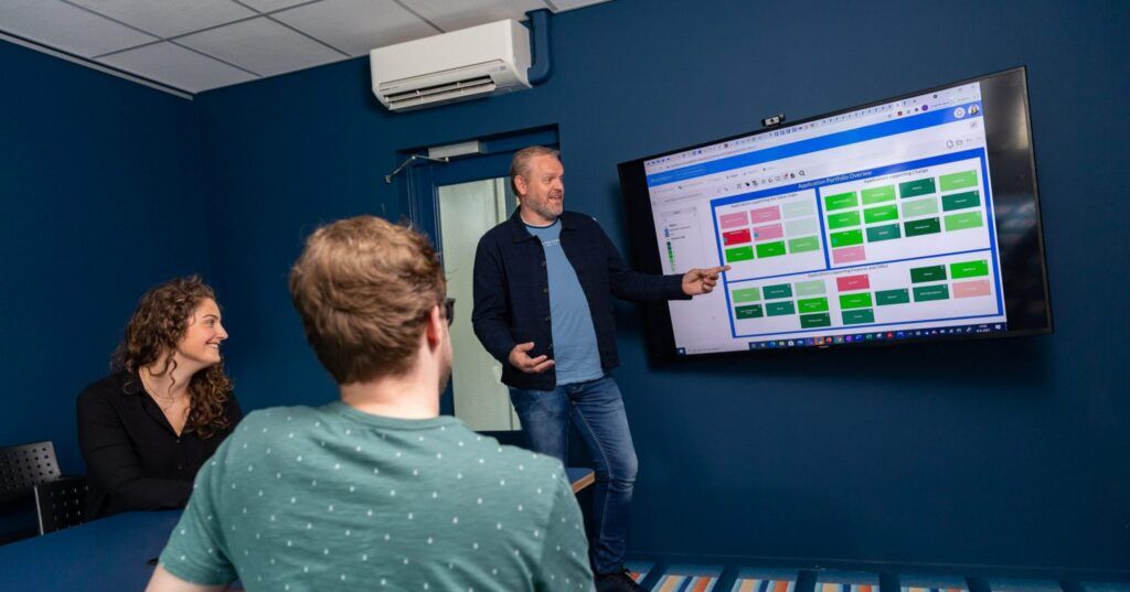 A man presents an application portfolio overview on a large screen to two colleagues in a meeting room.