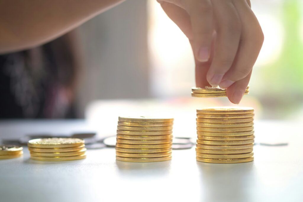 A hand placing a gold coin atop ascending stacks of gold coins on a desk, symbolizing growing returns.