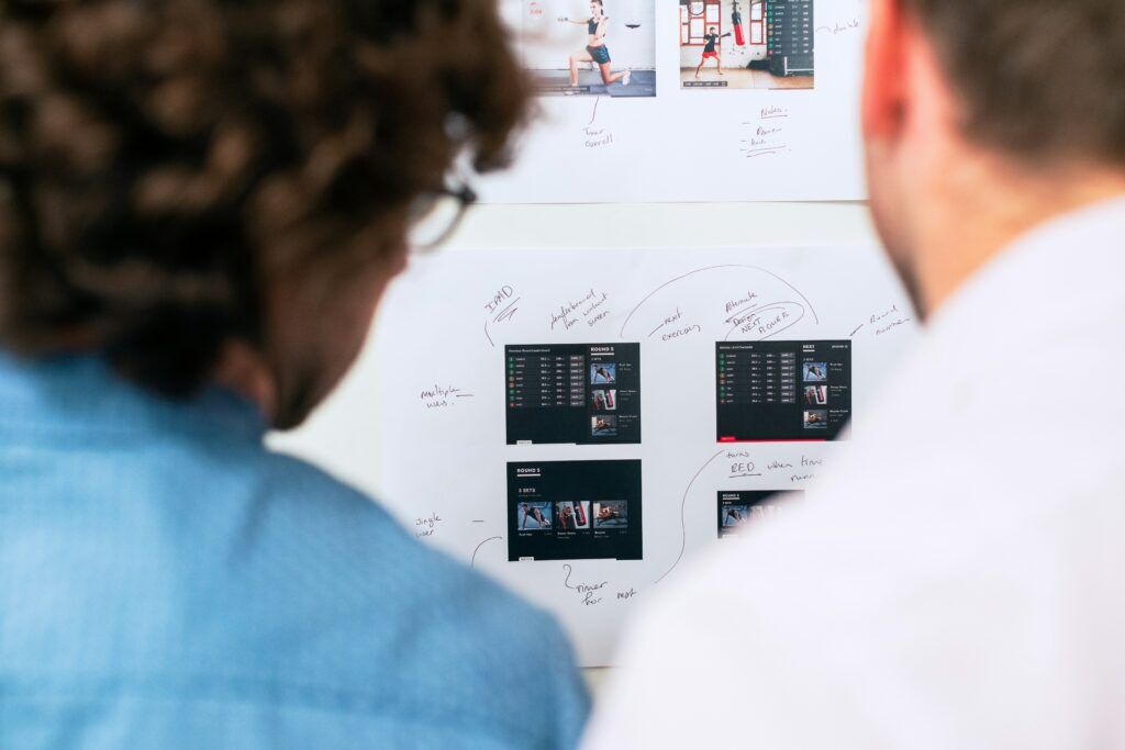 Two professionals reviewing a whiteboard with UI mockups, annotations, and design flow diagrams during a solution design