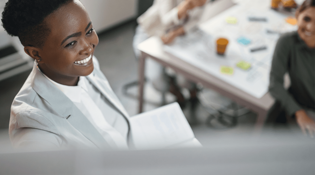 A smiling woman in a white blazer holds documents during a collaborative meeting with colleagues at a table.