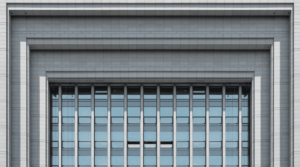 Symmetrical modern building facade with tall glass window panels framed by layered gray stone columns and horizontal ledges.