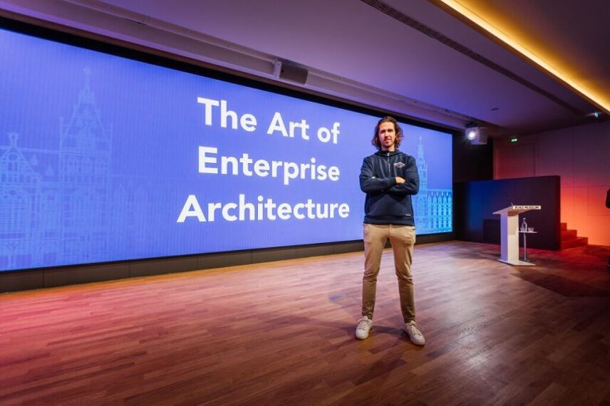 A man stands with arms crossed on a stage in front of a large screen reading "The Art of Enterprise Architecture."
