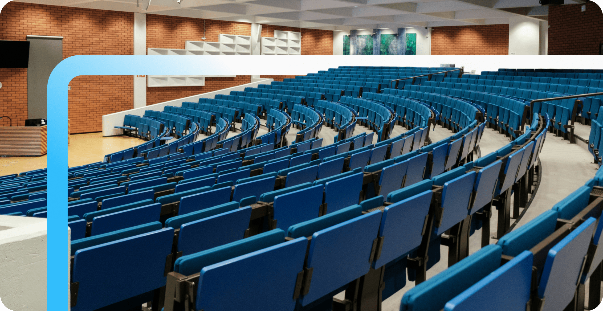 Empty university lecture hall with curved rows of blue tiered seating, a podium, and brick walls with acoustic panels.