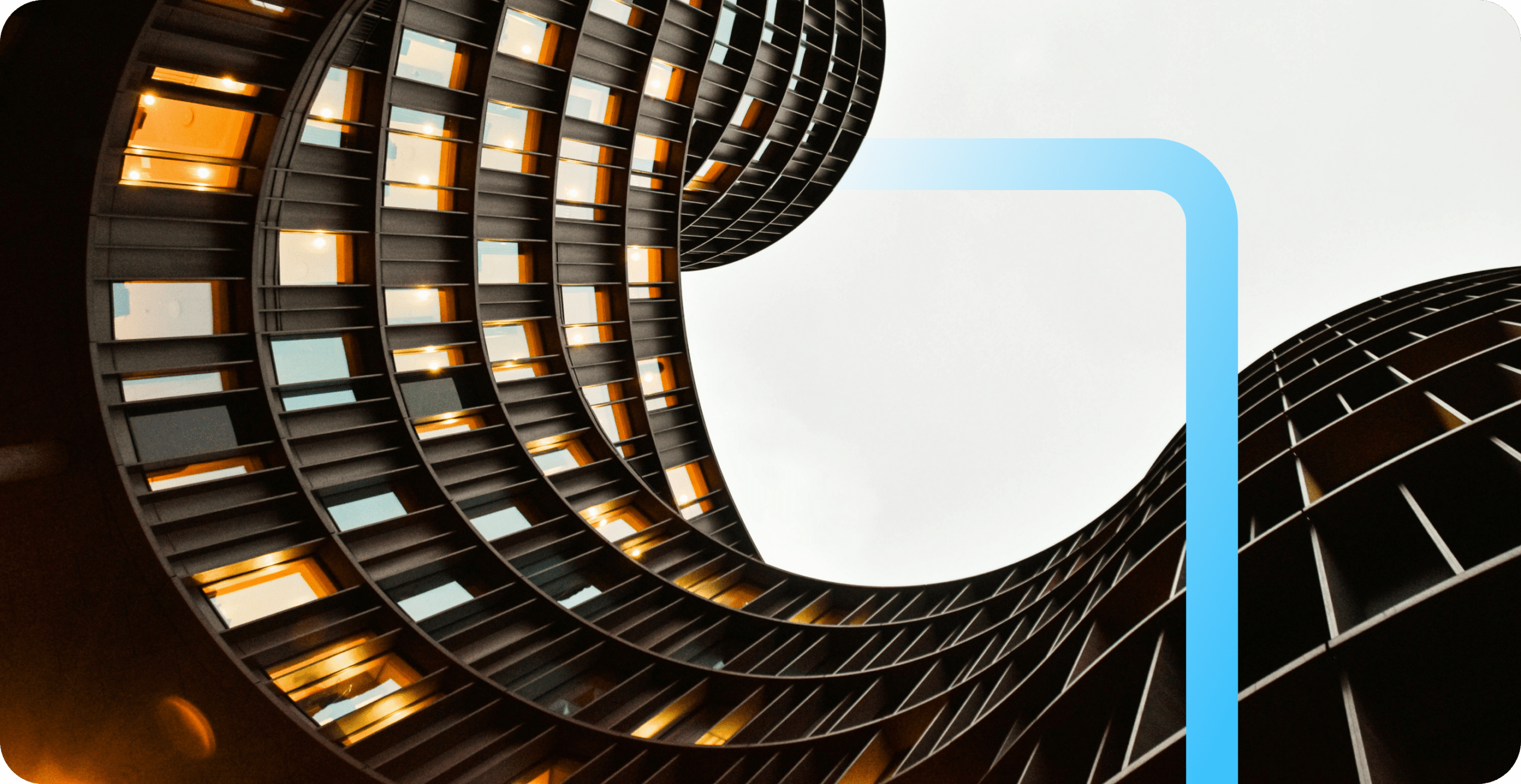 Upward view of curved modern buildings with warm-lit windows and a blue graphic overlay against a pale sky.