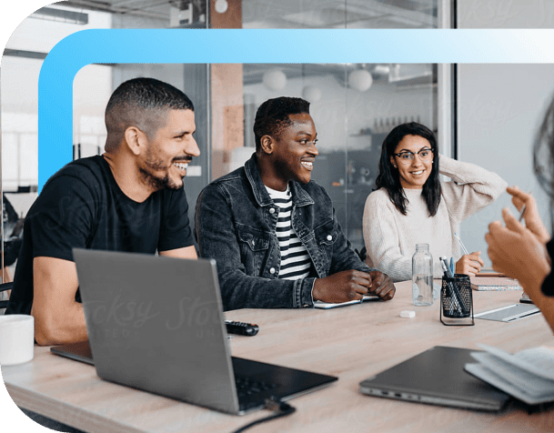 Smiling colleagues collaborating around a conference table with laptops in a modern glass-walled office space.
