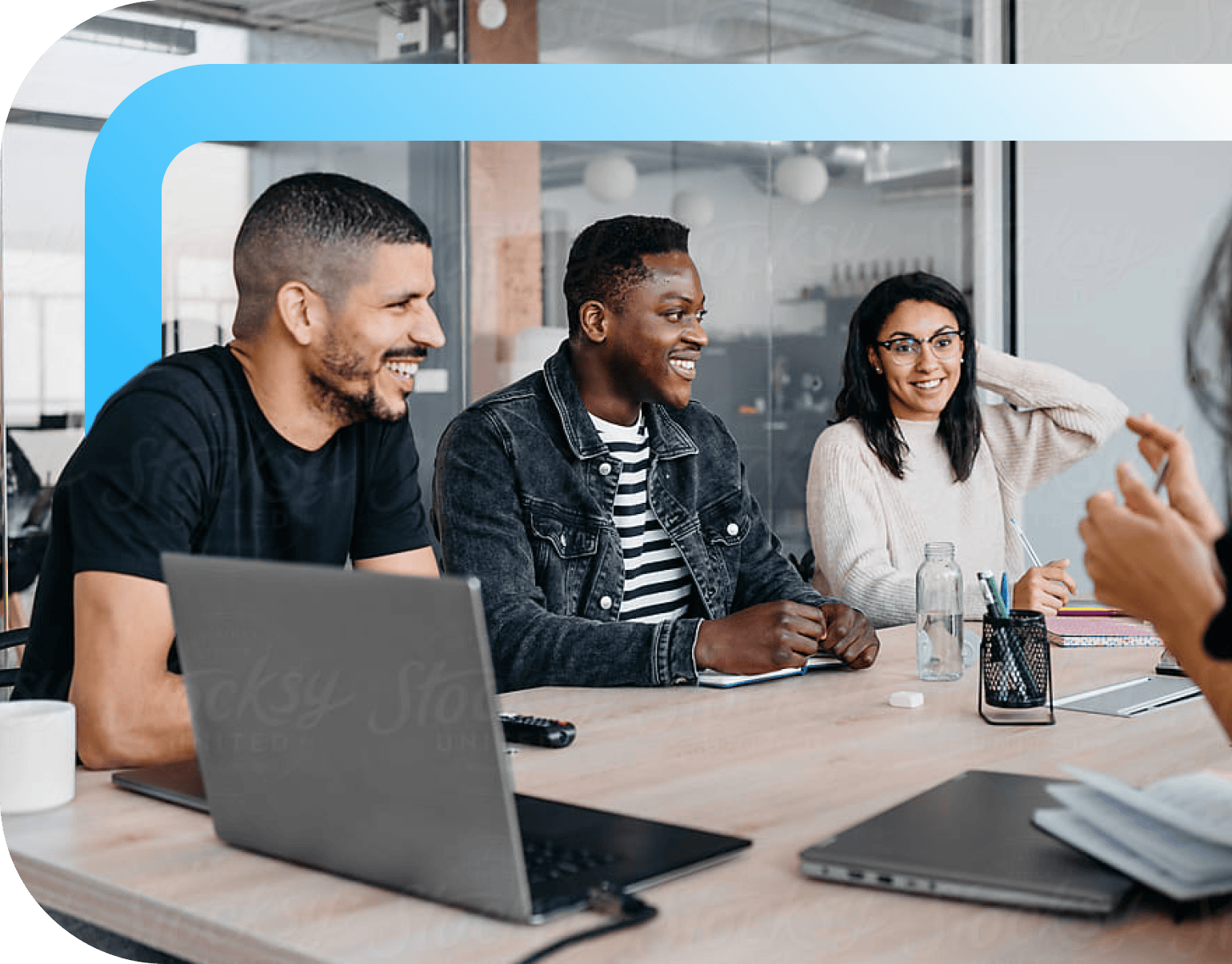 Smiling colleagues collaborating around a conference table with laptops in a modern glass-walled office space.