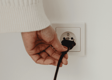 A hand plugs a black electrical cord into a white wall socket.