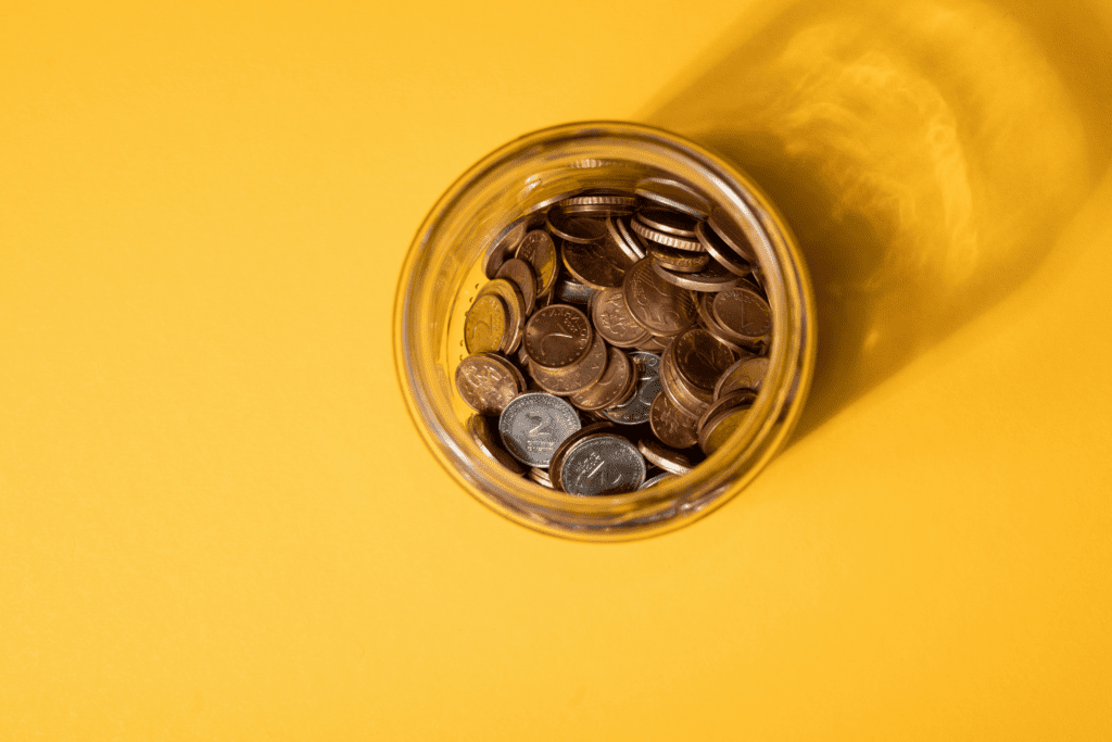 Glass jar filled with assorted coins viewed from above on a bright yellow background, symbolizing cost savings.