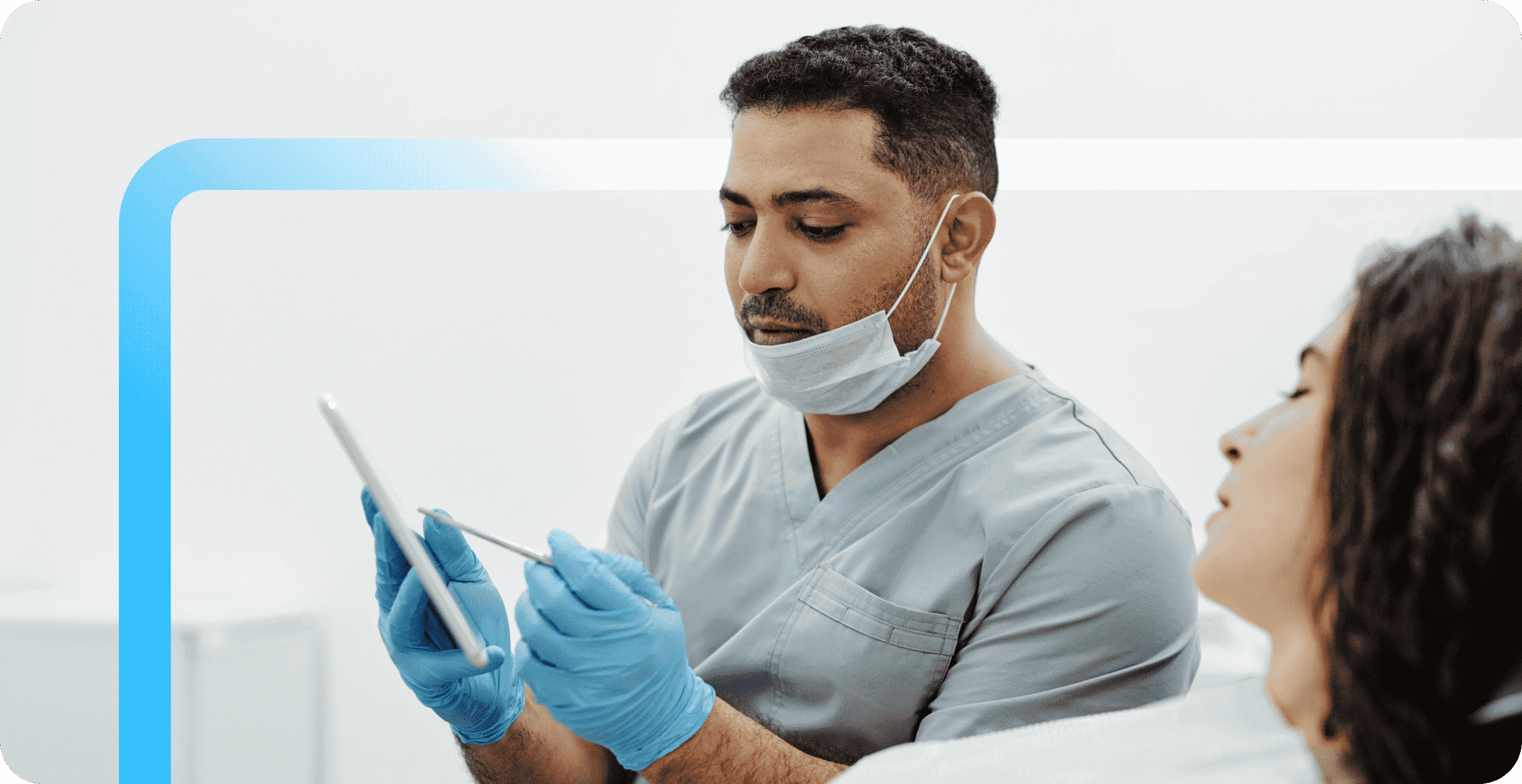 A dental professional in gray scrubs and blue gloves reviews a tablet while consulting with a female patient.